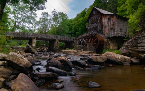 A rustic wooden mill with a water wheel beside a rocky stream and a bridge, surrounded by lush green trees.