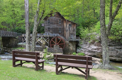 A rustic wooden mill with a water wheel, surrounded by lush greenery and two benches in the foreground.