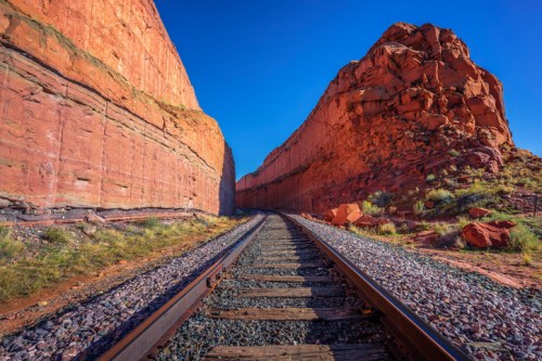 A railway track runs through a narrow canyon with towering red rock formations under a clear blue sky.