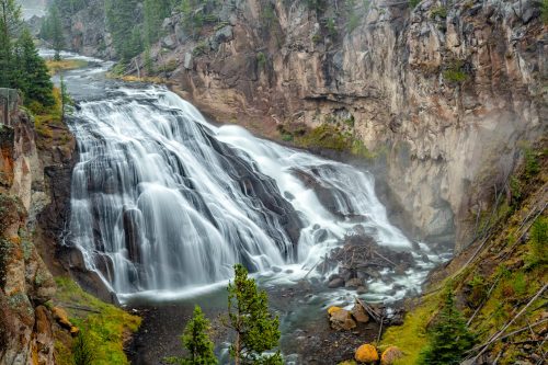 A cascading waterfall flows over rocky cliffs, surrounded by lush greenery and mist in a serene natural landscape.