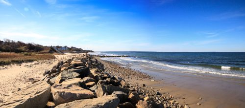 A panoramic view of a sandy beach with rocky shoreline, gentle waves, and clear blue skies.