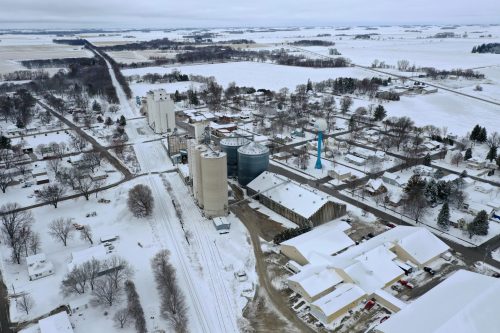 Aerial view of a snowy rural town with silos, buildings, and a road lined with trees in a winter landscape.
