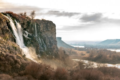 A scenic waterfall cascading down a rocky cliff, surrounded by barren trees and a calm river under a cloudy sky.
