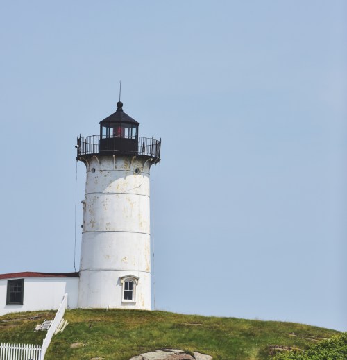 A weathered white lighthouse with a red roof stands on a grassy hill against a clear blue sky.