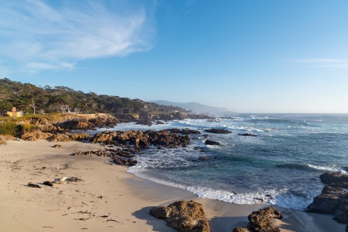 A serene beach scene with rocky shores, gentle waves, and a clear blue sky, surrounded by trees and distant hills.