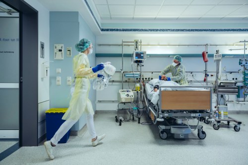 A healthcare worker in protective gear carries supplies in a hospital room while another attends to a patient.