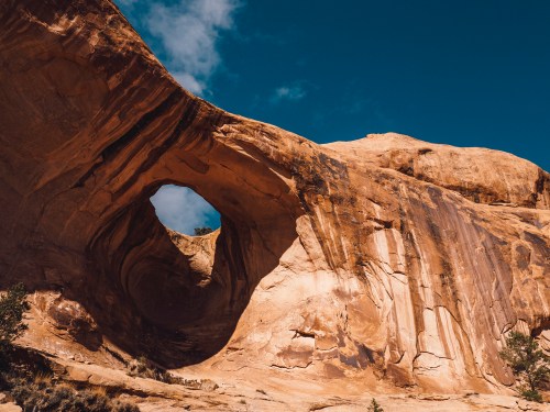 A large rock formation with a circular arch against a blue sky, showcasing textured sandstone and dramatic shadows.