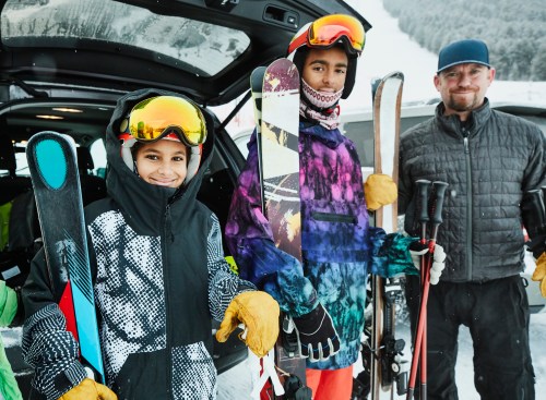 Two kids in ski gear hold skis and poles, smiling with a man in a winter jacket beside them, snowy landscape in the background.