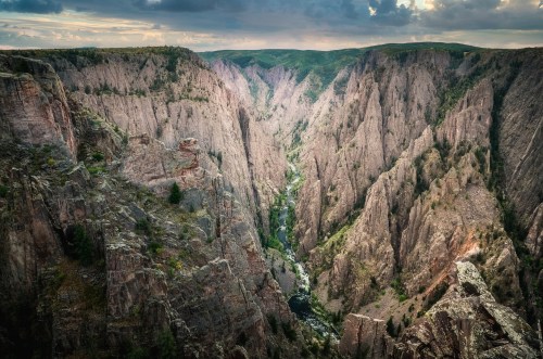 A dramatic canyon landscape with steep, rocky cliffs and a winding river below, under a cloudy sky.