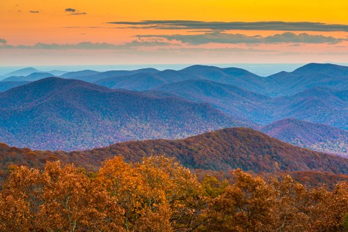 A panoramic view of rolling mountains at sunset, with vibrant autumn foliage in the foreground and soft, layered hills in the background.