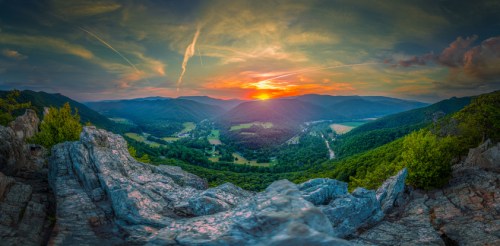 A panoramic view of a sunset over rolling hills and valleys, with rocky foreground and colorful sky.