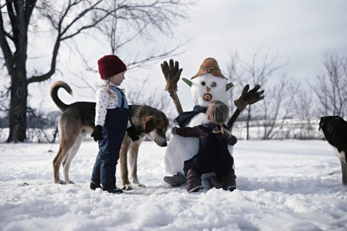 Two children and a dog play near a snowman in a snowy landscape, with trees in the background.
