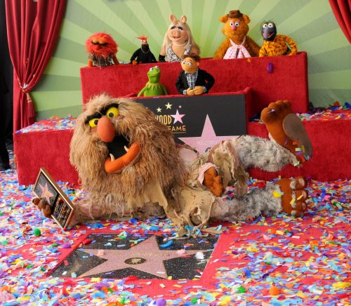 A Muppet character lounges on a star at the Hollywood Walk of Fame, surrounded by colorful confetti and other Muppets.