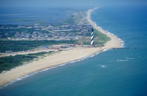 Aerial view of a coastal landscape featuring a striped lighthouse, sandy beach, and distant town along the shoreline.