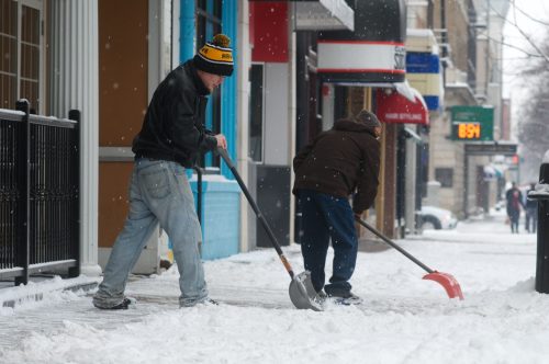 Two people shoveling snow from a sidewalk in a city, with shops visible in the background. Snow is falling.