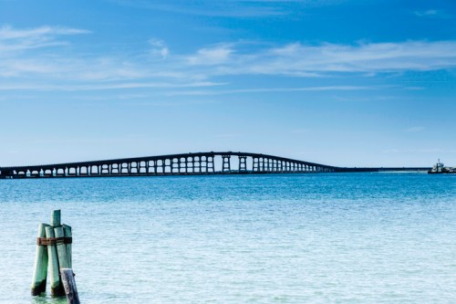 A long bridge spans across calm blue waters under a clear sky, with wooden posts in the foreground.