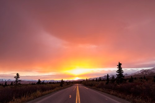 A scenic road stretches into the horizon under a vibrant sunset, with trees lining the sides and mountains in the background.
