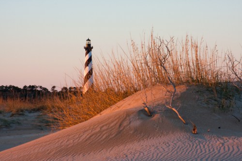 A striped lighthouse stands in the background, framed by sand dunes and tall grasses at sunset.