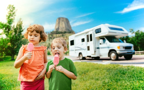 Two children enjoy popsicles in a grassy area with an RV and a mountain in the background on a sunny day.