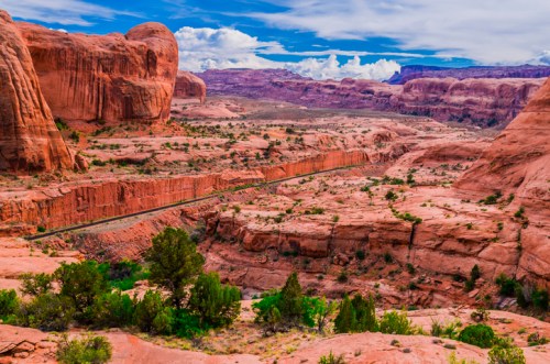 A scenic view of red rock formations and a winding road through a desert landscape under a blue sky with clouds.