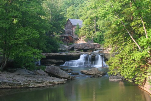 A serene landscape featuring a watermill beside a gentle waterfall, surrounded by lush green trees and rocky terrain.