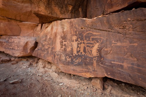 Ancient petroglyphs carved into a rock face, depicting various figures and symbols in a desert landscape.