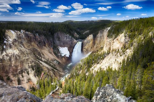 A stunning view of a waterfall cascading into a canyon, surrounded by lush green forests and a blue sky with clouds.