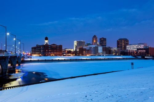 A winter cityscape at dusk, featuring a snowy riverbank and illuminated buildings in the background.