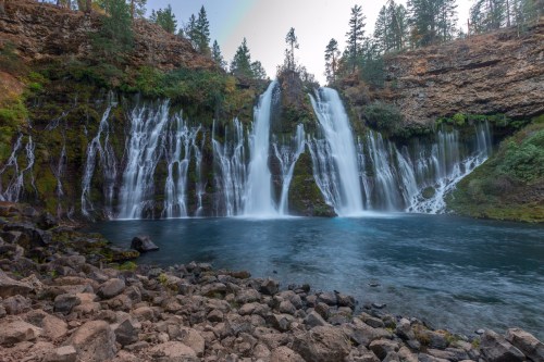 A serene waterfall cascades into a clear blue pool, surrounded by rocky terrain and lush green trees.