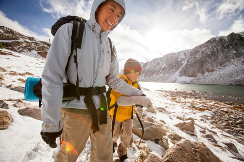A woman and child hike together in a snowy landscape, smiling with mountains and a lake in the background.