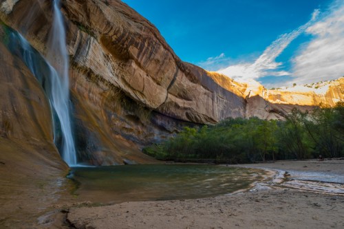 A serene waterfall cascades down a rocky cliff into a calm pool, surrounded by lush greenery and blue skies.