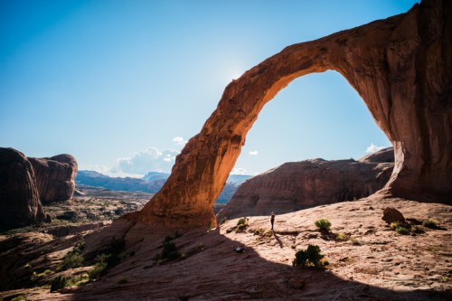 A natural stone arch stands against a clear blue sky, with a person exploring the rocky landscape below.