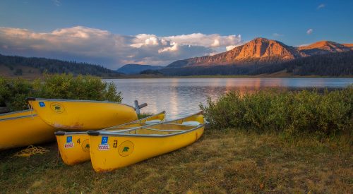 Three yellow canoes by a serene lake, surrounded by lush greenery and mountains under a colorful sunset sky.