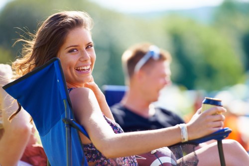 A young woman smiles while sitting in a chair, holding a drink, with a blurred figure in the background.