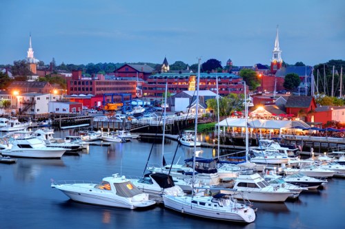 A scenic harbor at dusk, filled with boats, surrounded by colorful buildings and church steeples in the background.