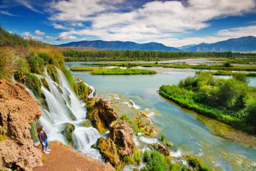 A person stands near a waterfall, surrounded by lush greenery and mountains under a blue sky with fluffy clouds.