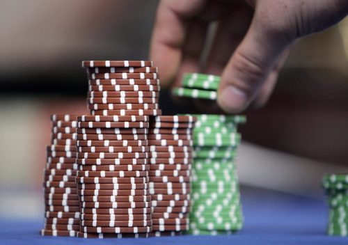 A hand stacking poker chips on a blue table, with tall towers of brown and green chips in the foreground.