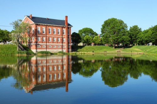 A red brick building reflects in a calm lake, surrounded by green trees and clear blue skies.