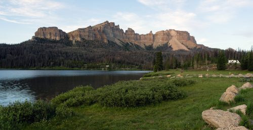 A serene lake reflects towering mountains, surrounded by lush greenery and rocky shores under a clear sky.