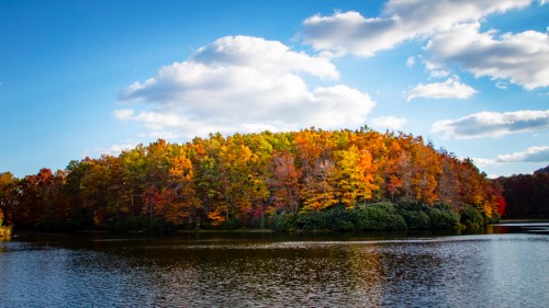 A serene lake surrounded by vibrant autumn foliage under a blue sky with fluffy clouds.