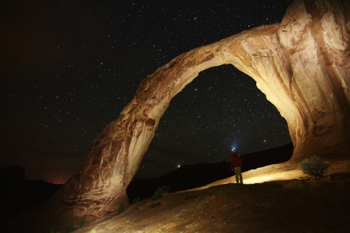 A person stands beneath a large rock arch at night, illuminated by stars and a flashlight, creating a serene atmosphere.