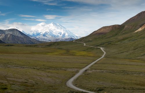 A winding road leads through a vast green valley, with snow-capped mountains in the background under a blue sky.