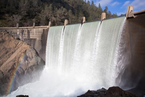 A dam with multiple spillways releasing water, creating a mist and a faint rainbow in the foreground.