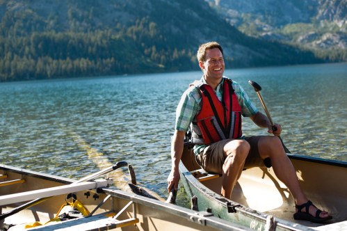 A smiling man in a life jacket sits in a canoe on a calm lake, surrounded by mountains and trees.