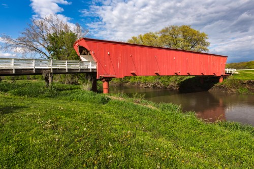 A red covered bridge spans a river, surrounded by green grass and trees under a partly cloudy sky.