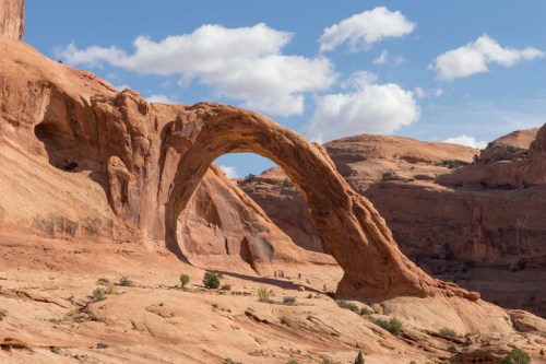 Natural rock arch in a desert landscape, surrounded by red sandstone formations and a blue sky with clouds.