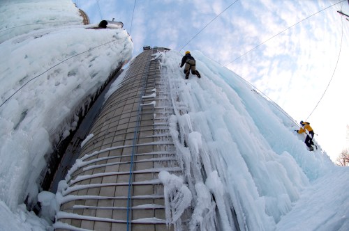 Two climbers ascend icy structures, with one on a ladder and the other scaling a frozen wall under a cloudy sky.