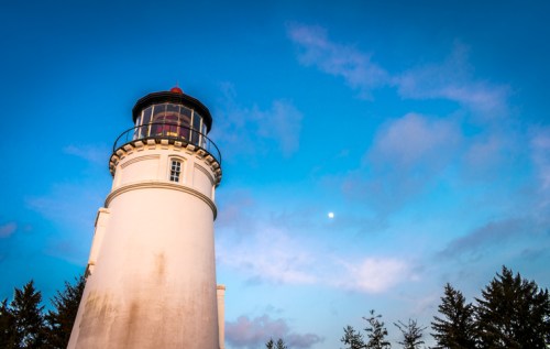 A tall lighthouse against a blue sky with clouds and a visible moon, surrounded by trees.