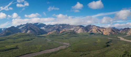 A panoramic view of colorful mountains under a blue sky with scattered clouds, surrounded by lush green valleys.