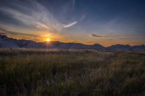 Sunset over a rugged landscape with rolling hills and tall grass, casting warm light across the scene.